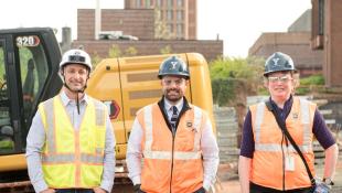 Three construction people in orange and yellow vests standing in front of a crane