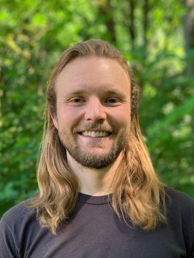 Headshot of Jackson Cooper in a brown t-shirt with green foliage in the background