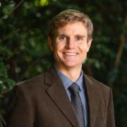 Headshot of Matthew Browning in a gray blazer, blue shirt and tie, and green foliage in the background