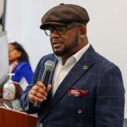Headshot of Charles T. Brown in a blue suit and brown cap standing with a microphone