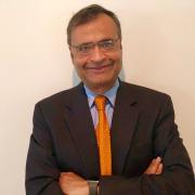 Headshot of Anadi Jauhari in dark suit and orange tie against white wall