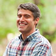 Headshot of Greg Bratman in plaid shirt with foliage in the background