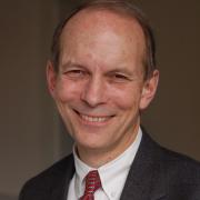 Headshot of Paul Cleary in red tie and dark suit against brown background